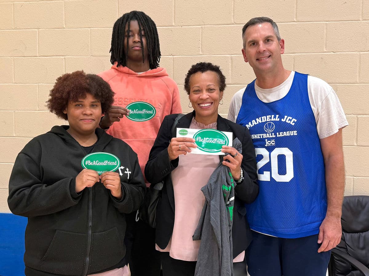 Four people posing together indoors, three of them holding green #BeAGoodFriend stickers