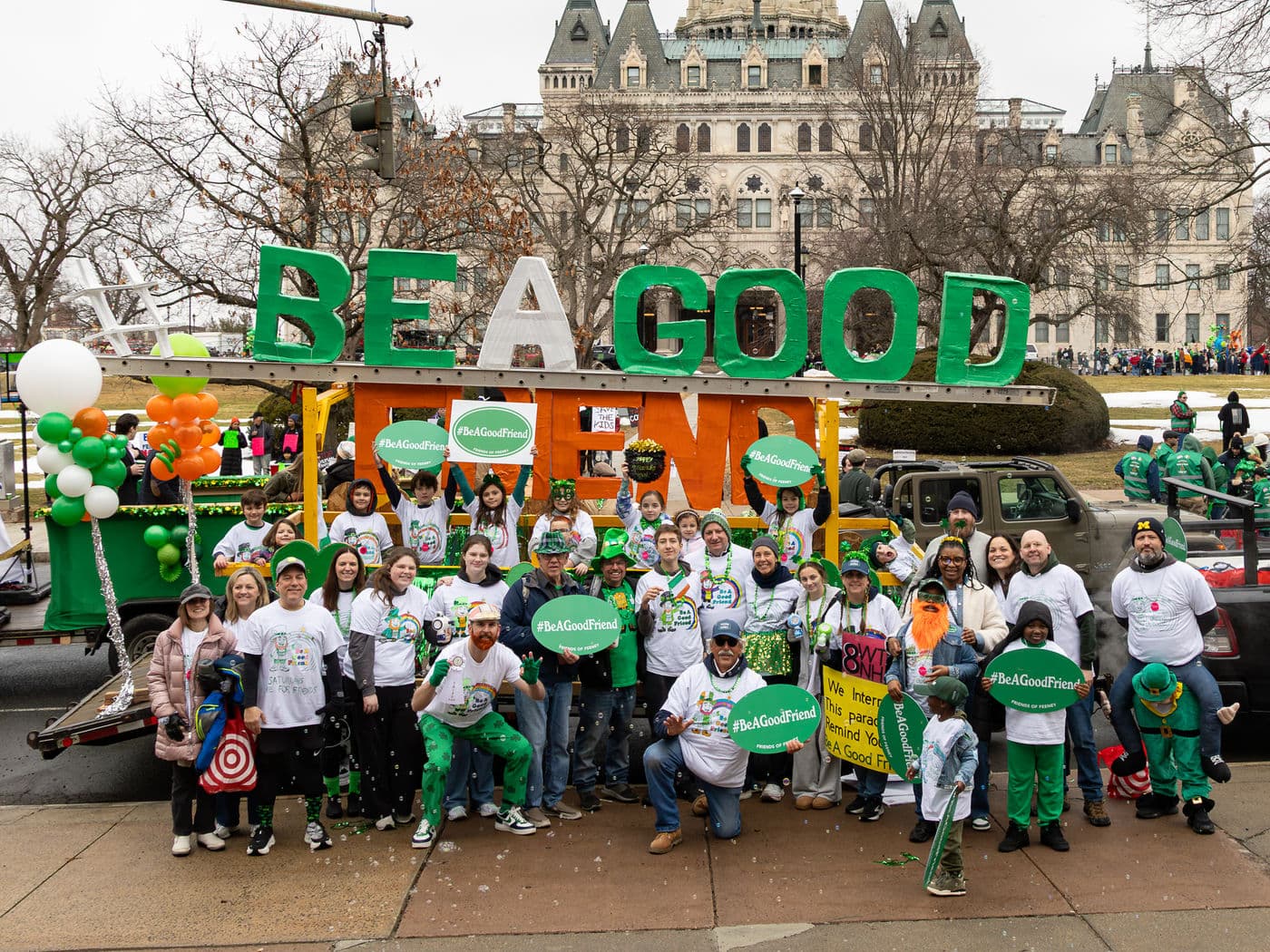 Marchers in white #BeAGoodFriend shirts and green hats in front of the BE A GOOD FRIEND float