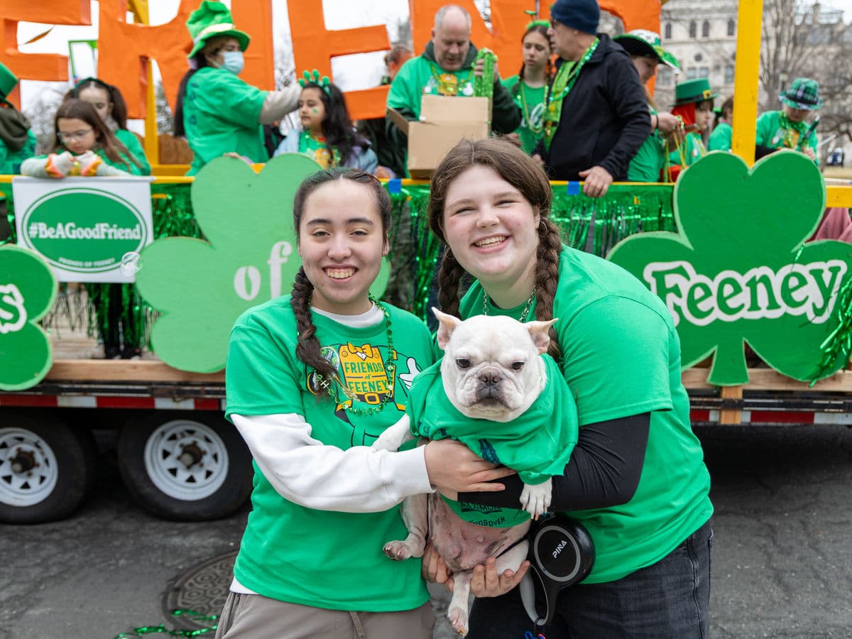 Two marchers in green shirts holding a bulldog in front of the shamrock-covered float