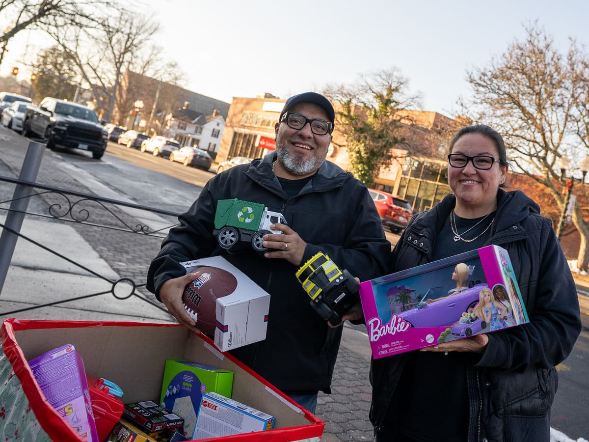 Two people holding donated toys beside a full collection bin