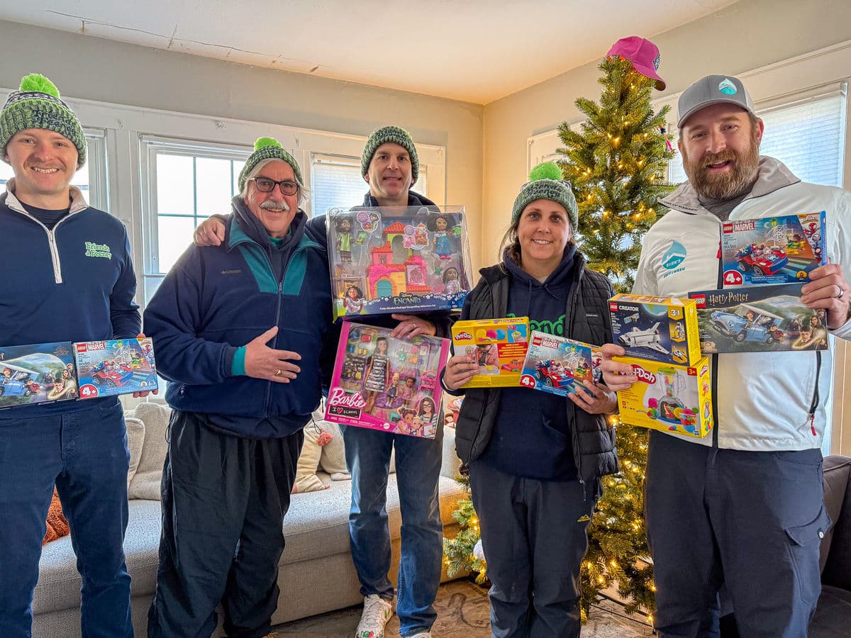 Volunteers in green knit hats holding boxed toys in front of a Christmas tree