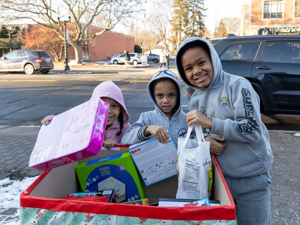 Three children in hoodies holding gifts beside a toy collection box