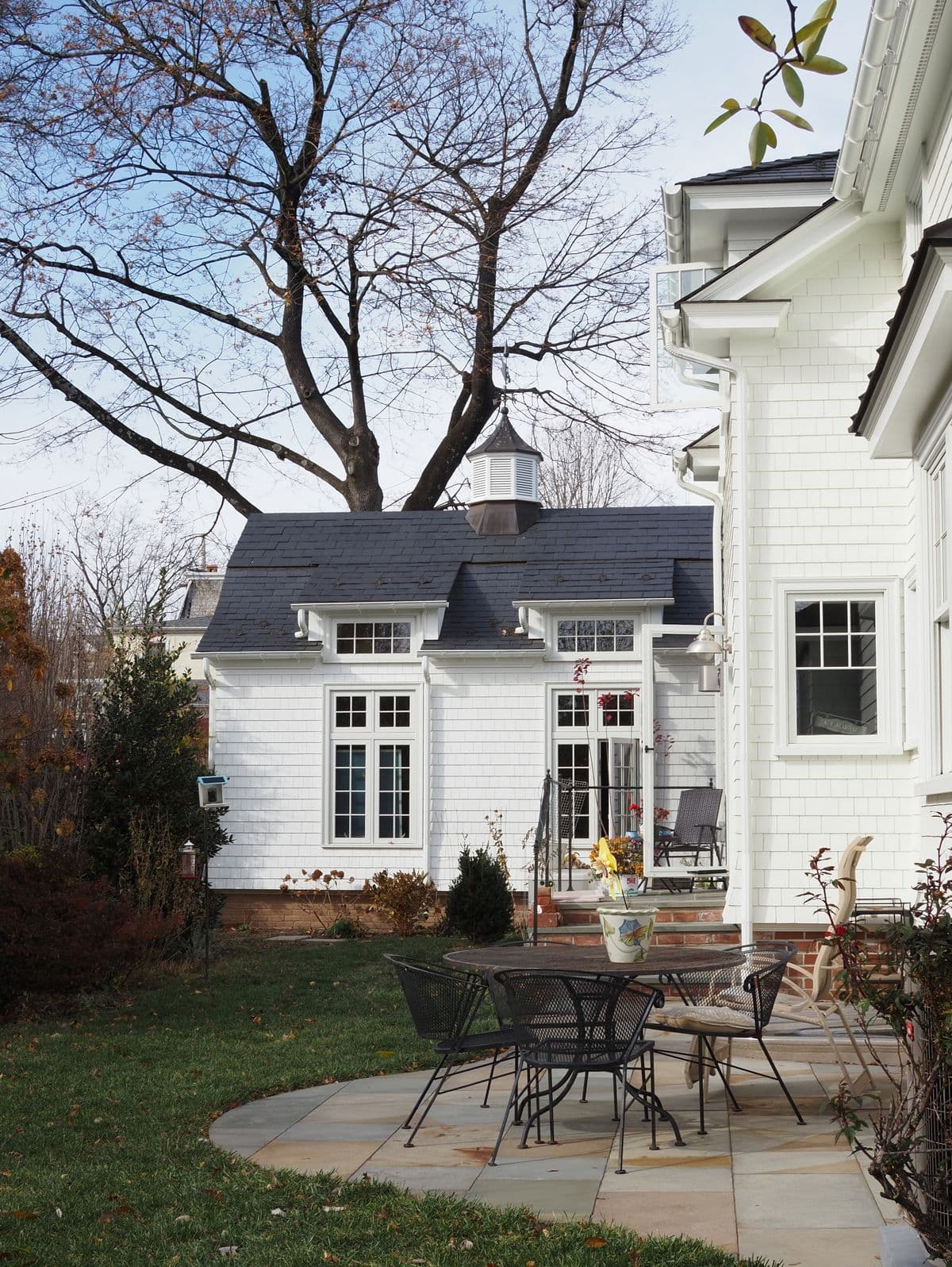 A curved bluestone patio with a wrought-iron table set beside a white shingled house and carriage house in late fall