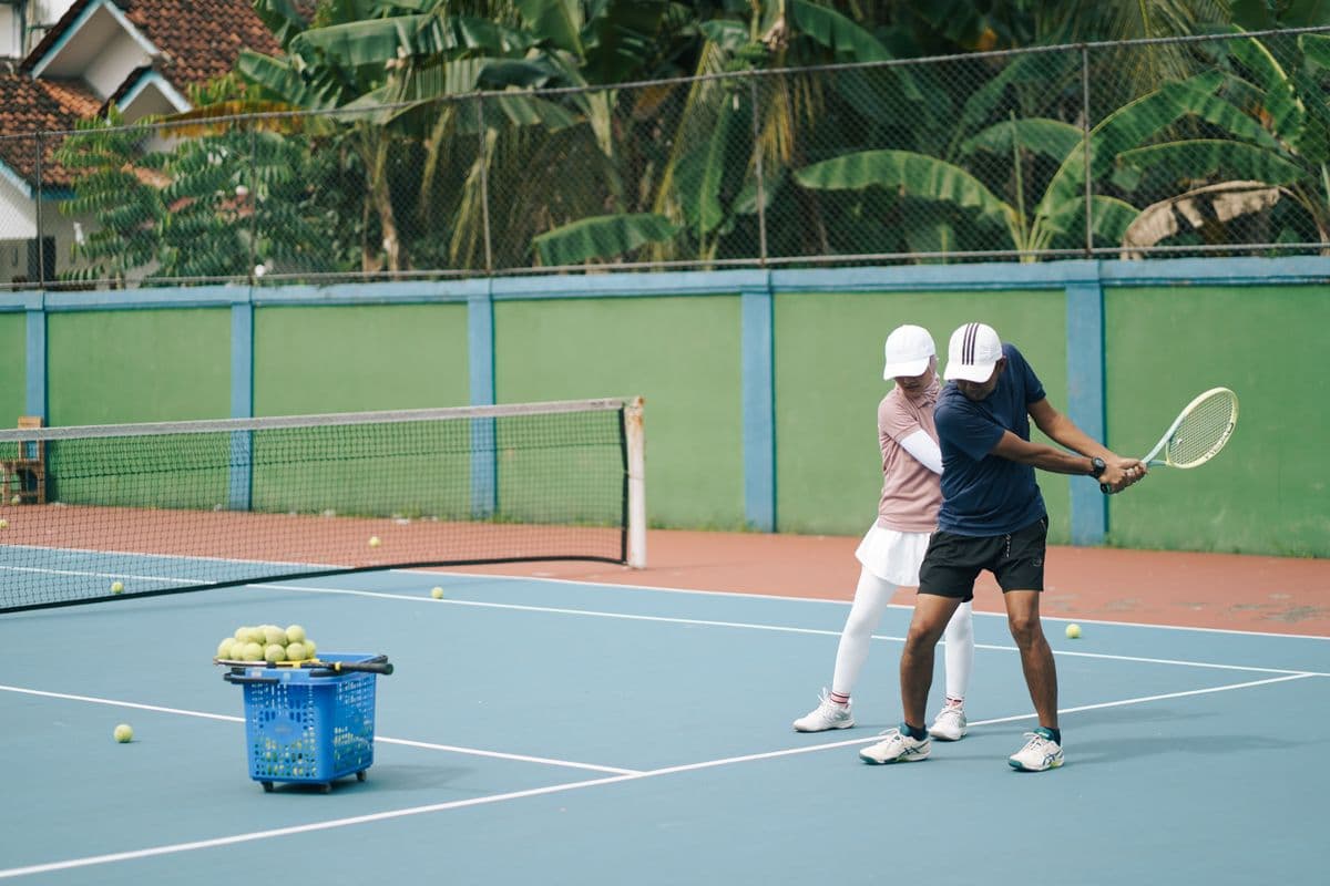 A coach guides a player through a backhand swing next to a basket of balls