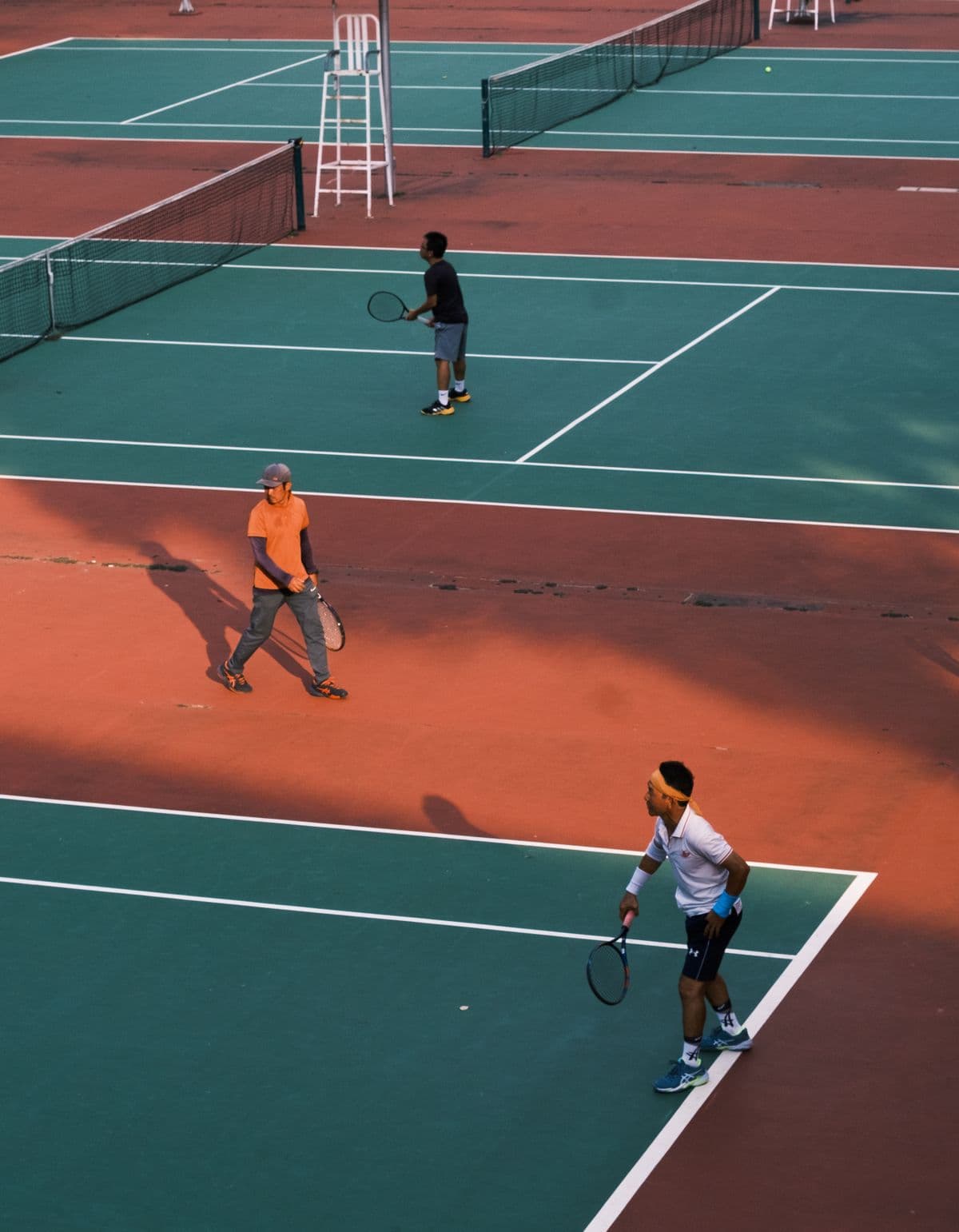 Adults playing doubles on green courts with red surrounds, seen from above