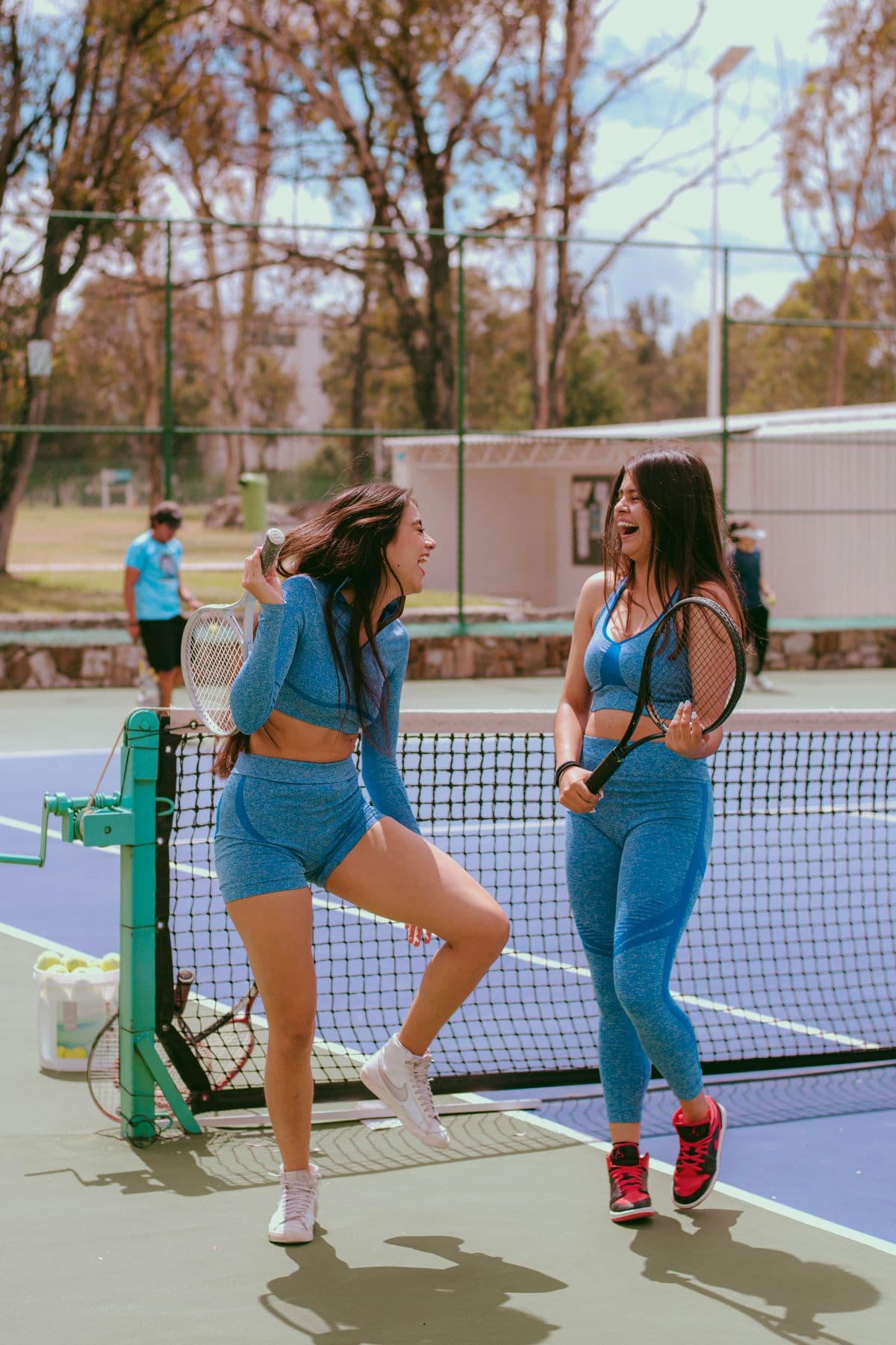 Two women in matching blue sportswear laugh together at the net between points