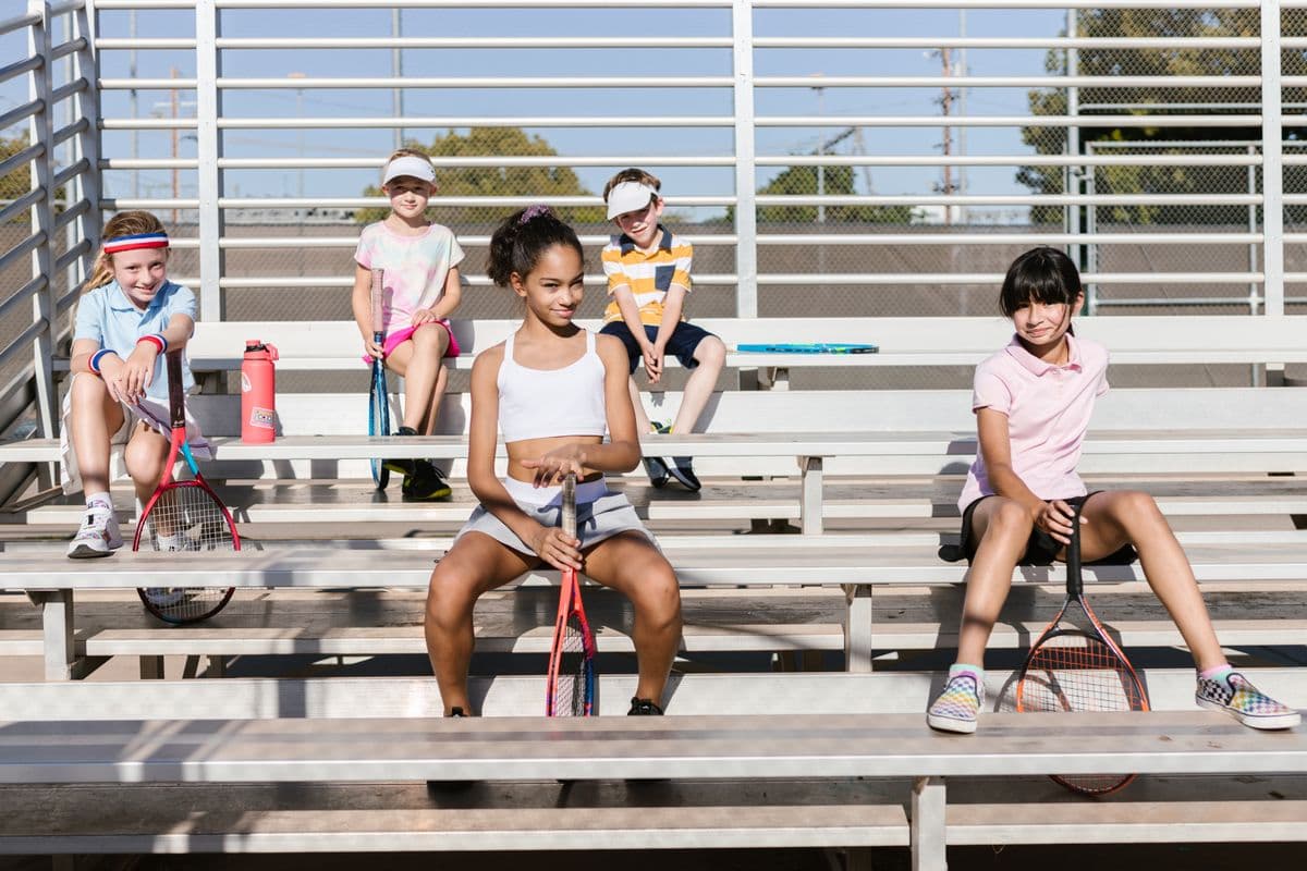 Five campers sit on courtside bleachers with their rackets on a sunny morning