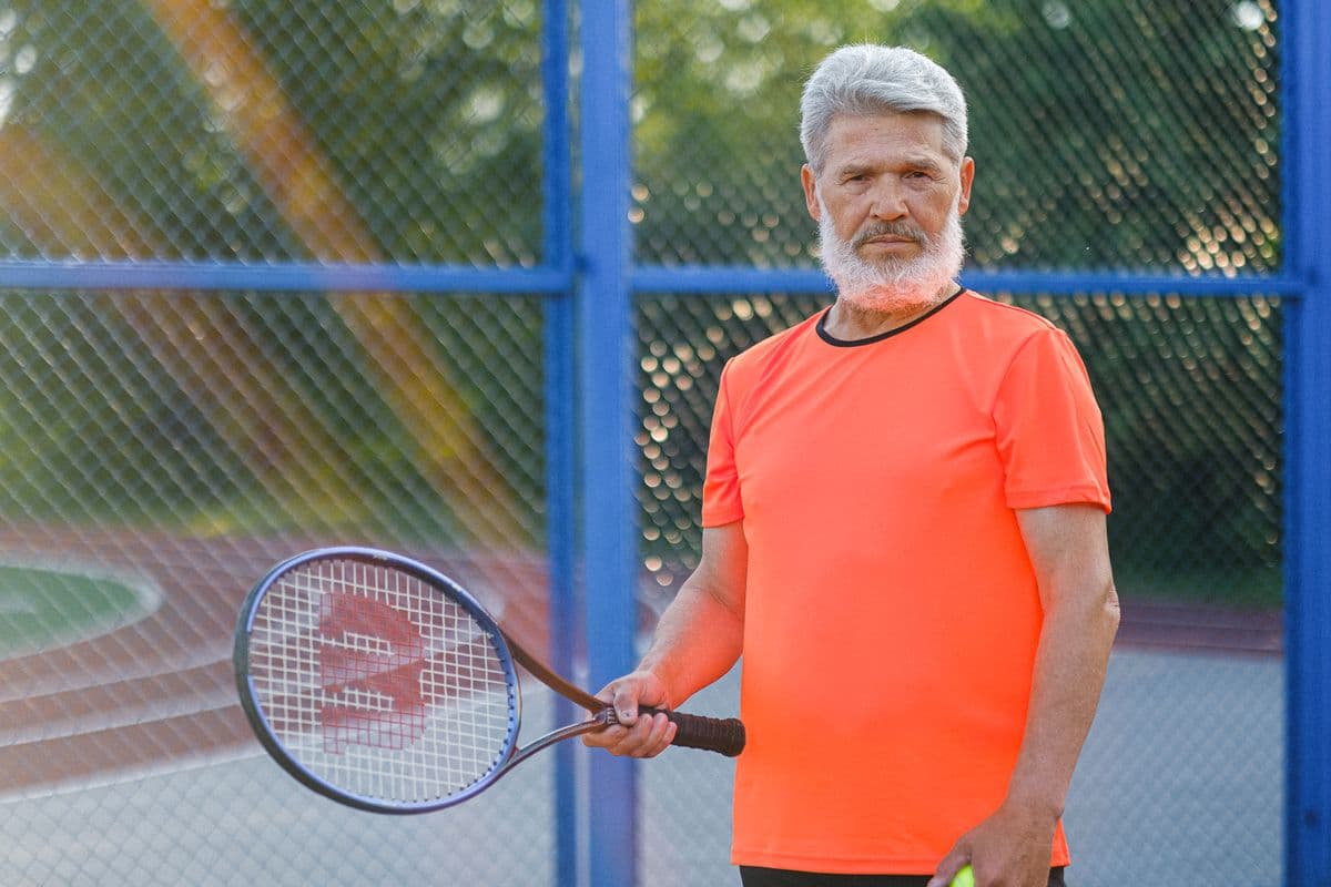 Dan Whitaker, a gray-bearded coach in an orange shirt, holding a racket by a blue fence