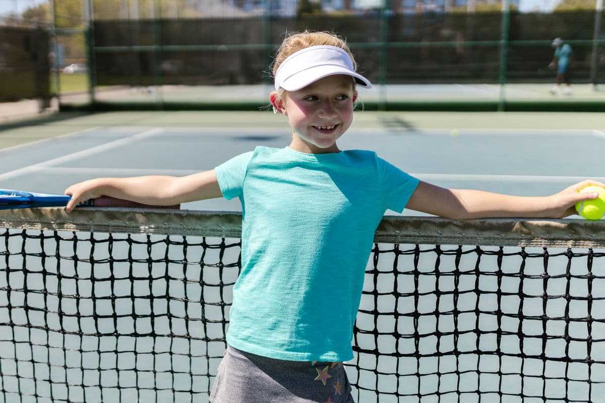 A smiling girl in a white visor leans on the net with a racket in one hand and a ball in the other