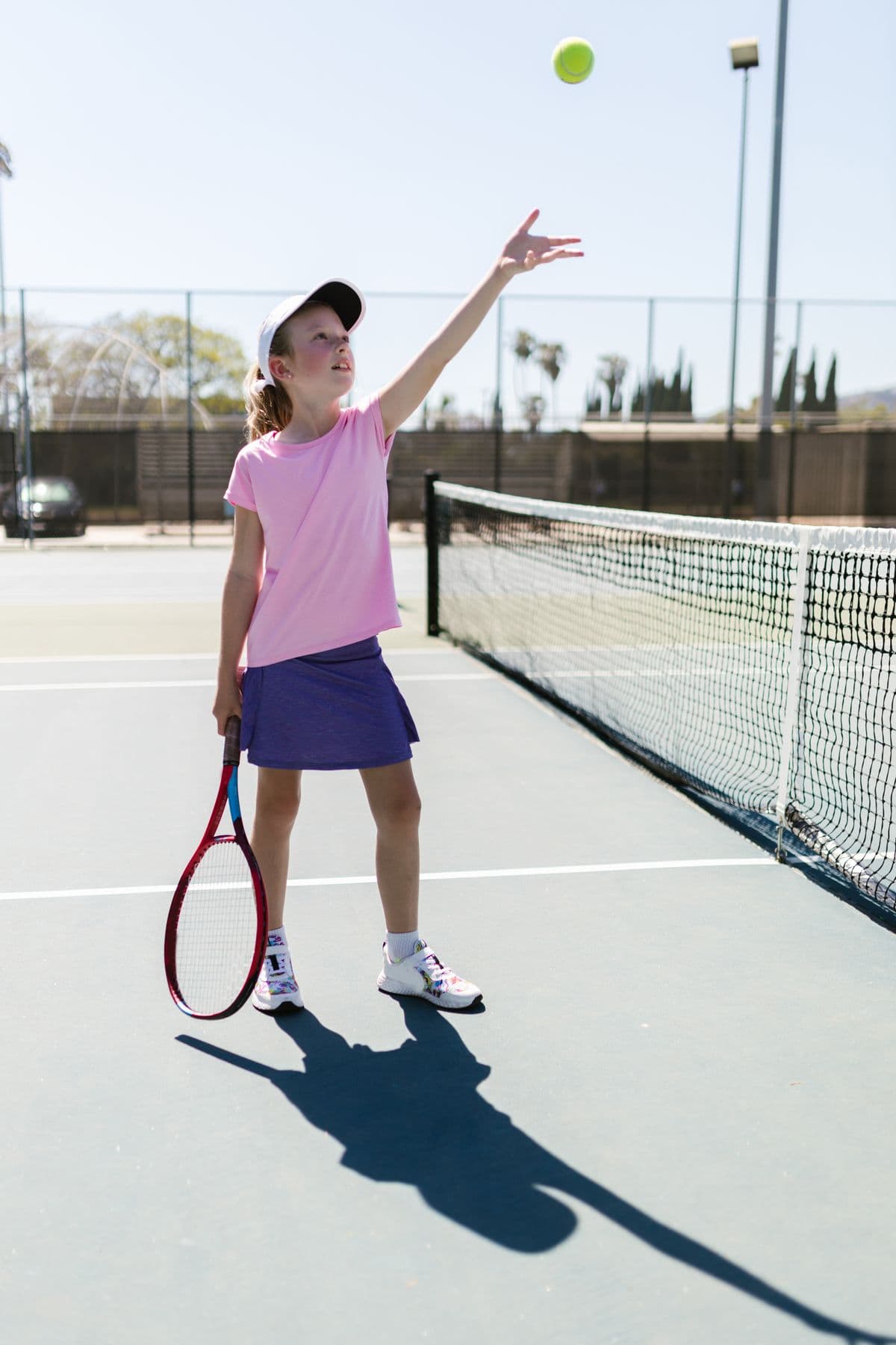 A girl in a pink shirt tosses the ball up to serve beside the net