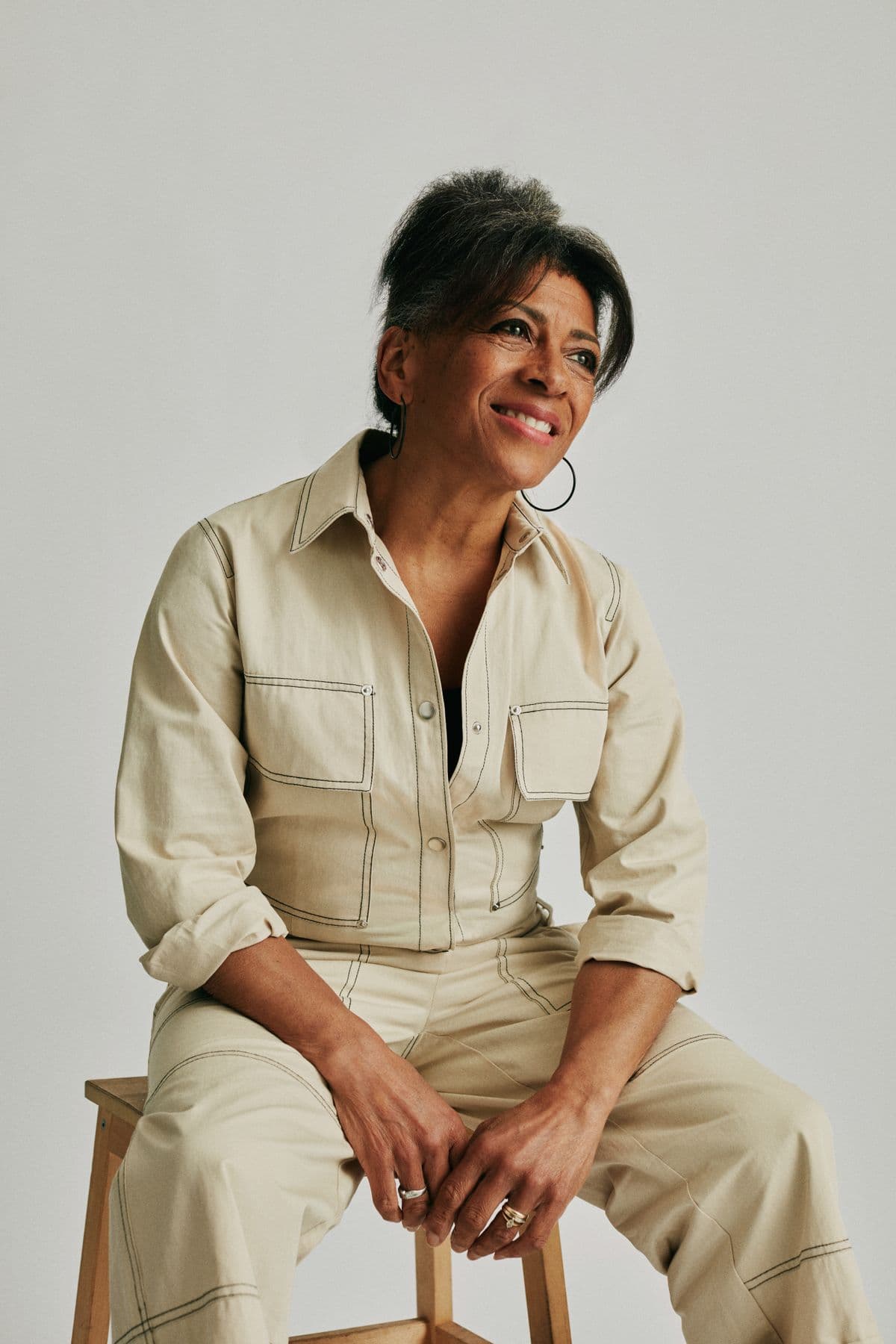 Dana Whitfield smiling, seated on a wooden stool in a cream jumpsuit