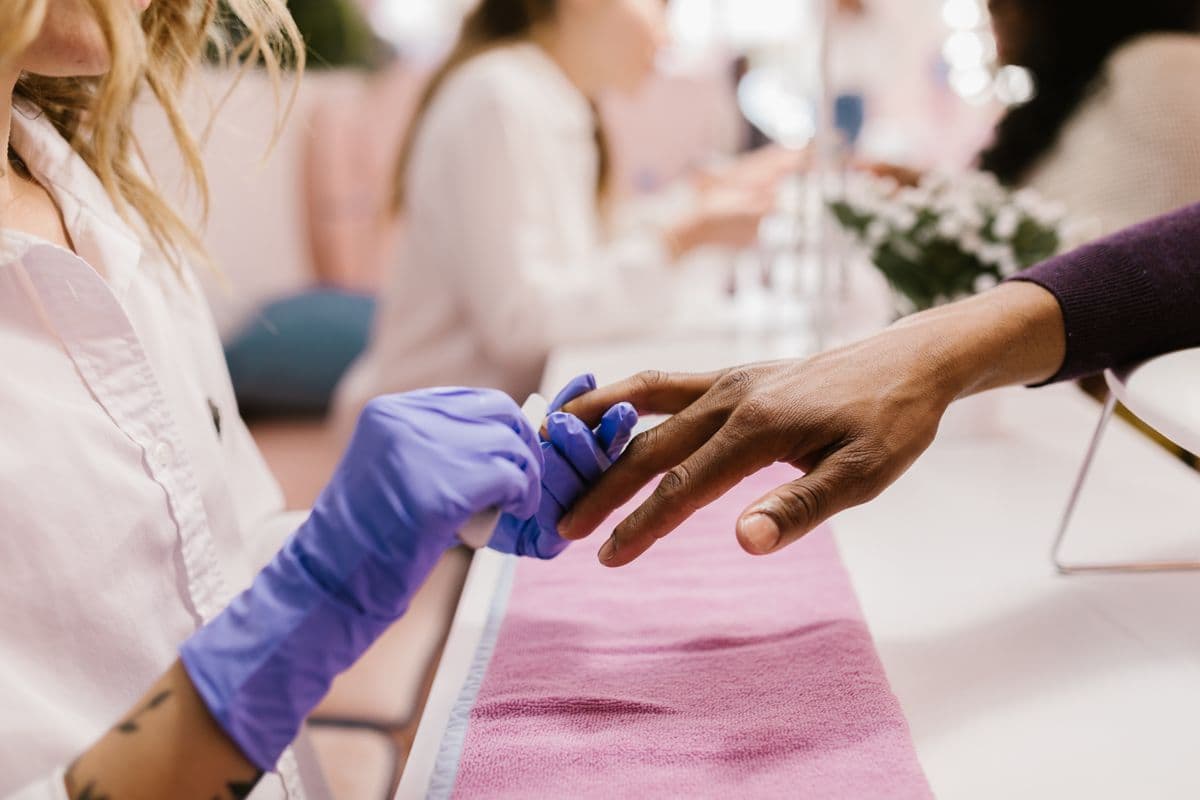 A nail artist in lavender gloves shaping a client’s nails at a white manicure table
