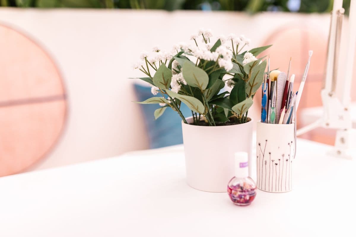 A white manicure table with a potted plant, a cup of fine brushes and a bottle of cuticle oil