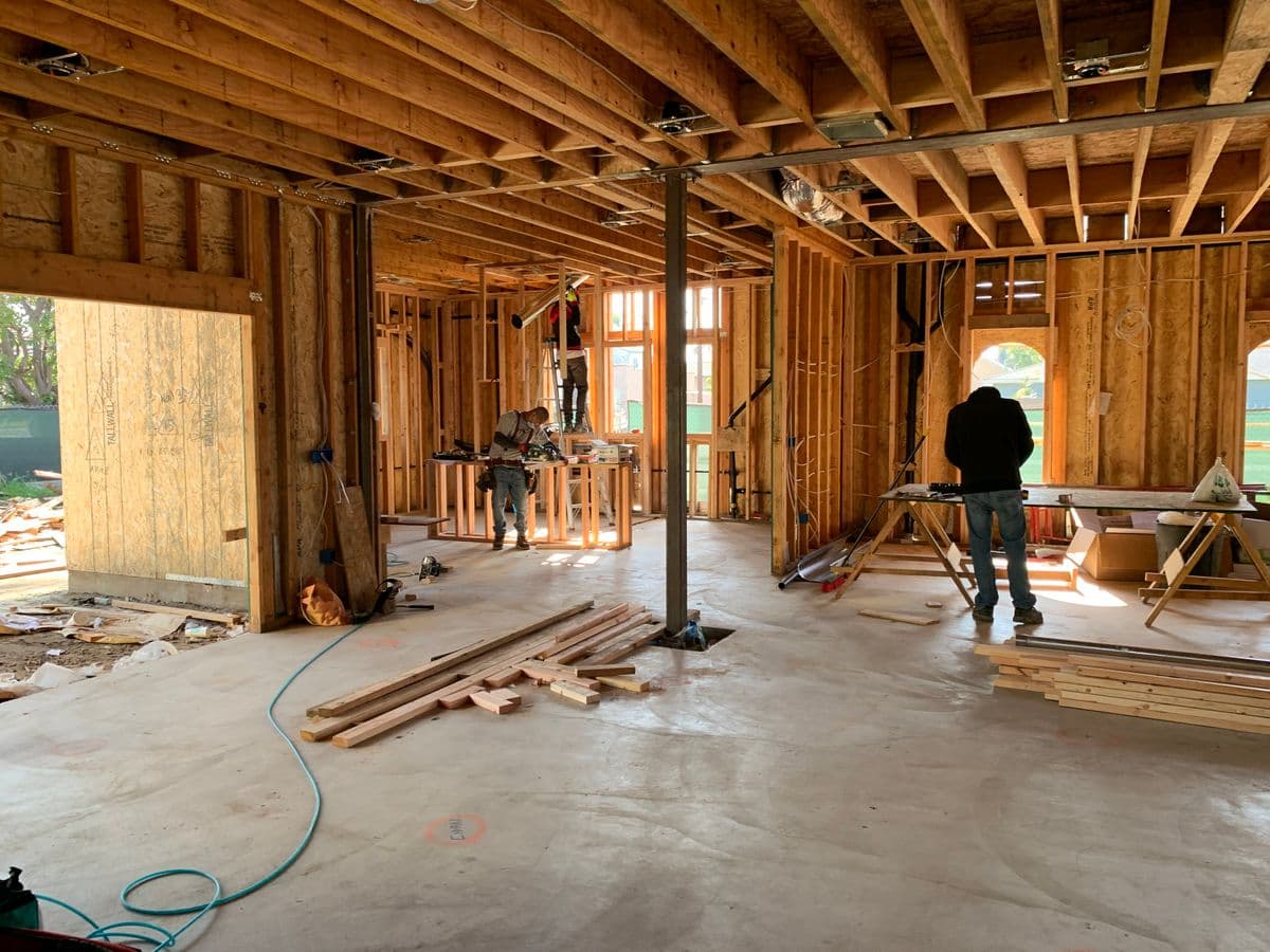 Crew working inside new stud walls and floor joists before sheathing, lumber stacked on the slab