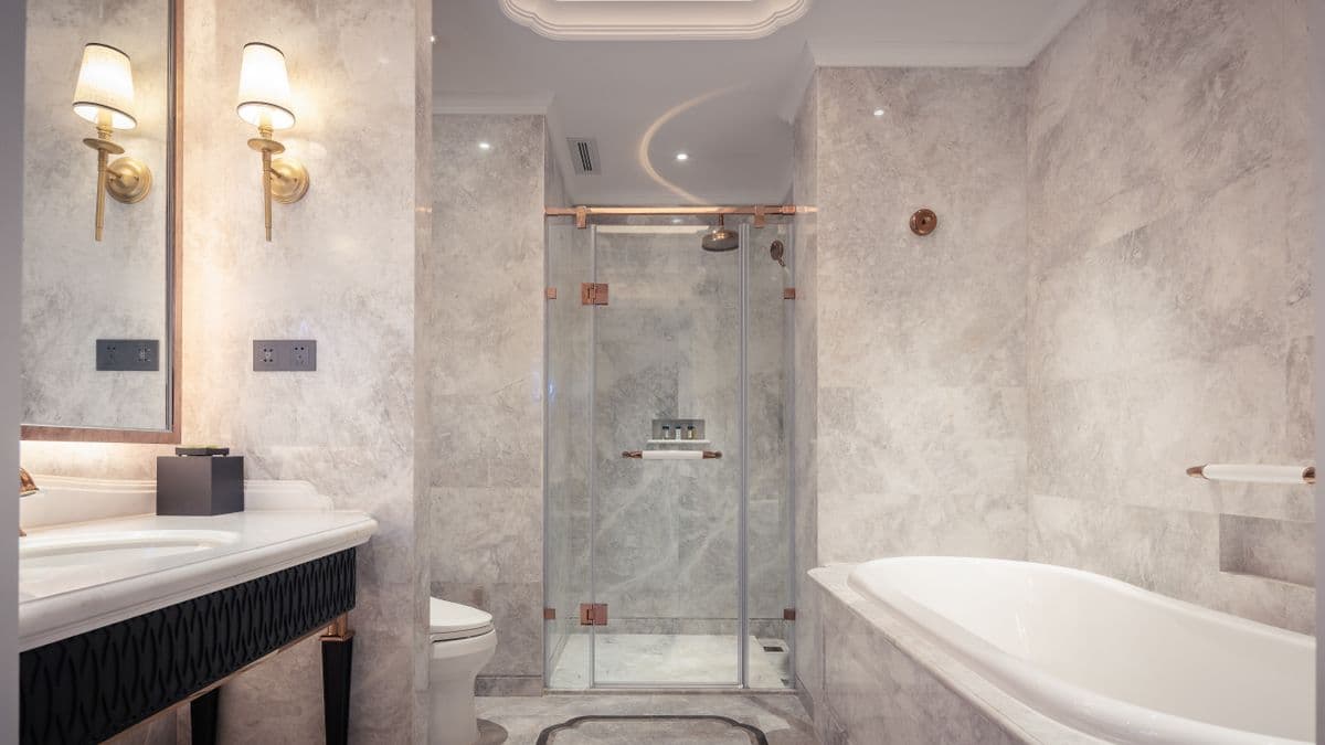 Glass walk-in shower beside a tub, with gray marble walls and brass sconces
