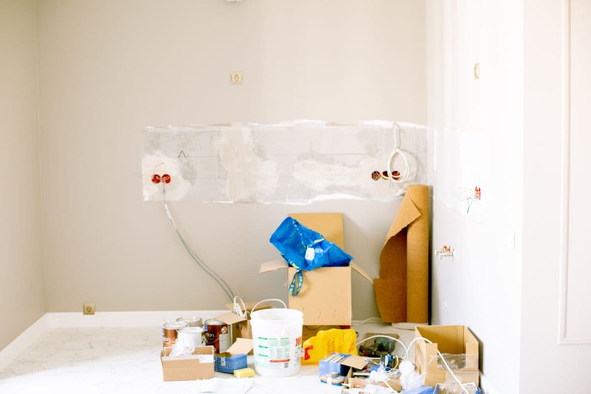 Kitchen wall stripped to the plaster mid-demolition, with capped plumbing and boxes of debris on the counter