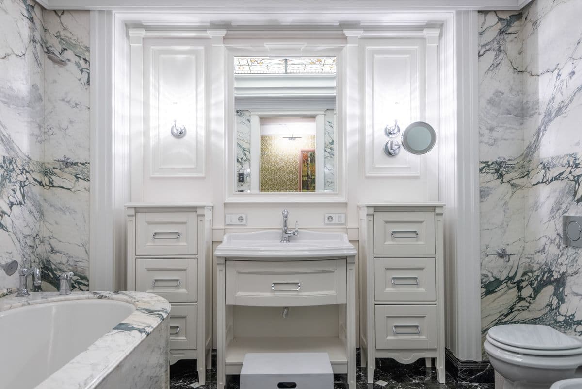 Marble bathroom with a white paneled vanity, wall sconces and a tub with a marble surround