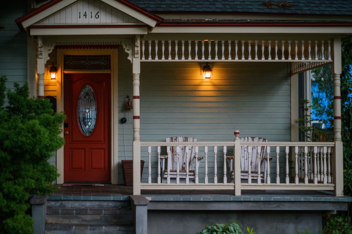 Covered front porch with a white railing, wooden chairs, porch lights and a red front door