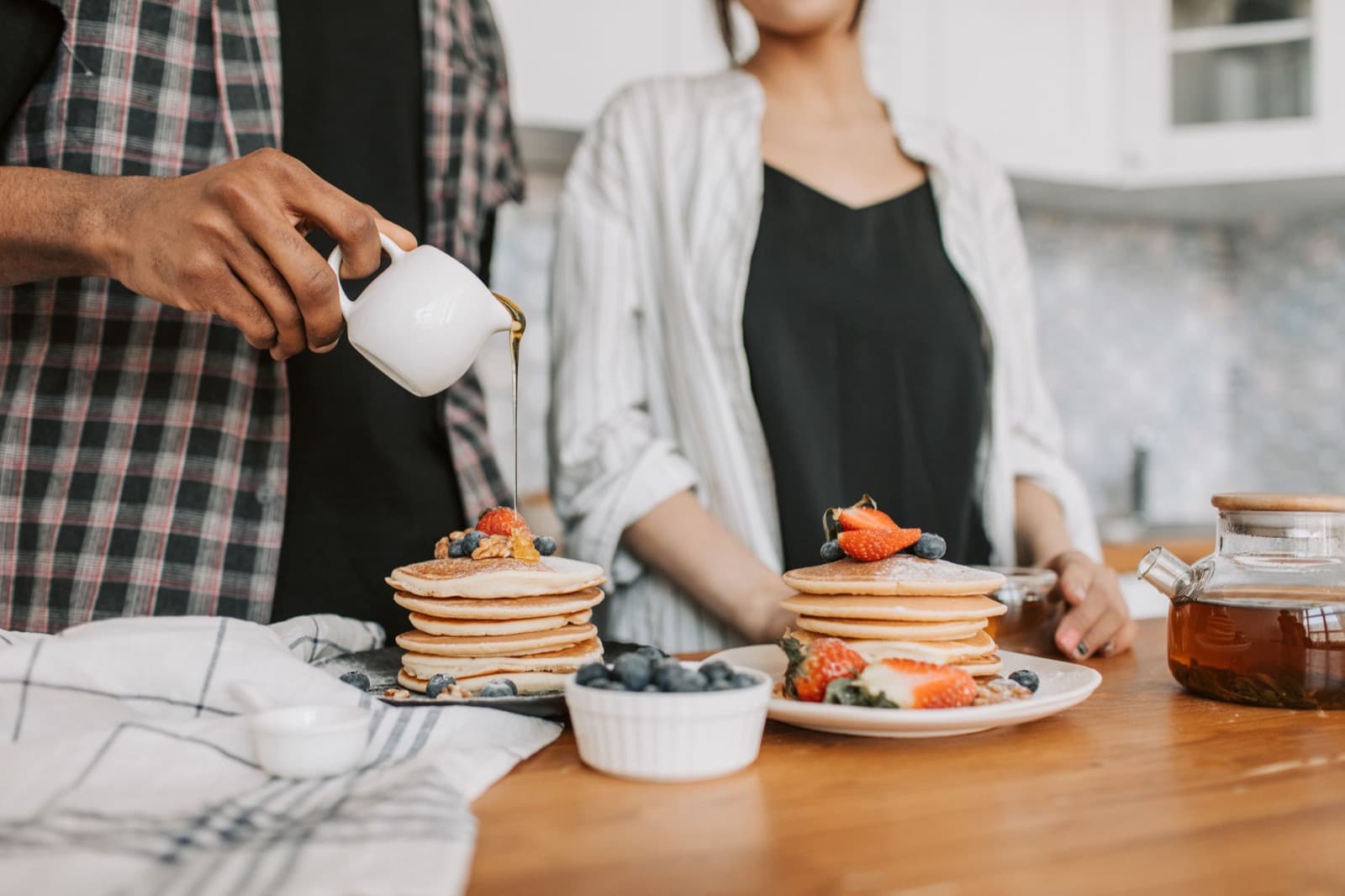 Maple syrup poured from a small jug over a stack of pancakes topped with berries