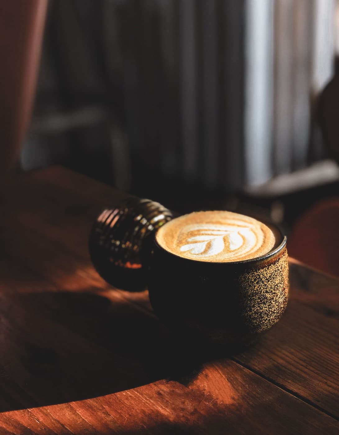 A cortado in a speckled stoneware cup catching window light on a dark wood table