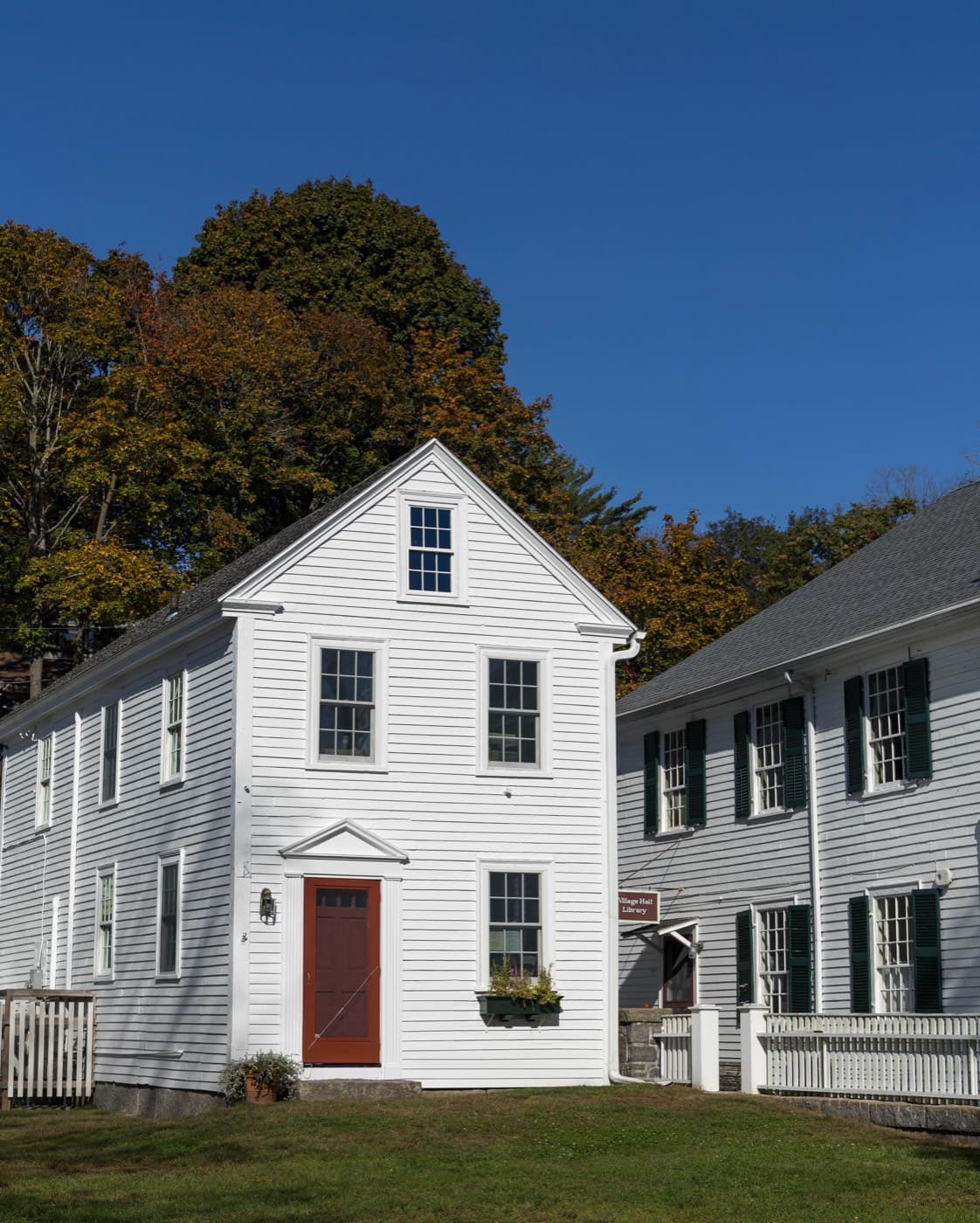 A white clapboard colonial house with black shutters and a red front door under a clear autumn sky