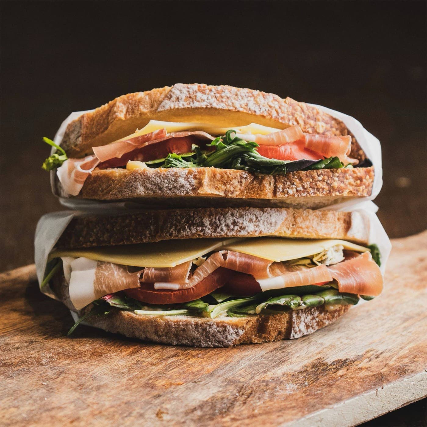 A tall sourdough sandwich with cheese, greens and cured meat, halved on a wooden board