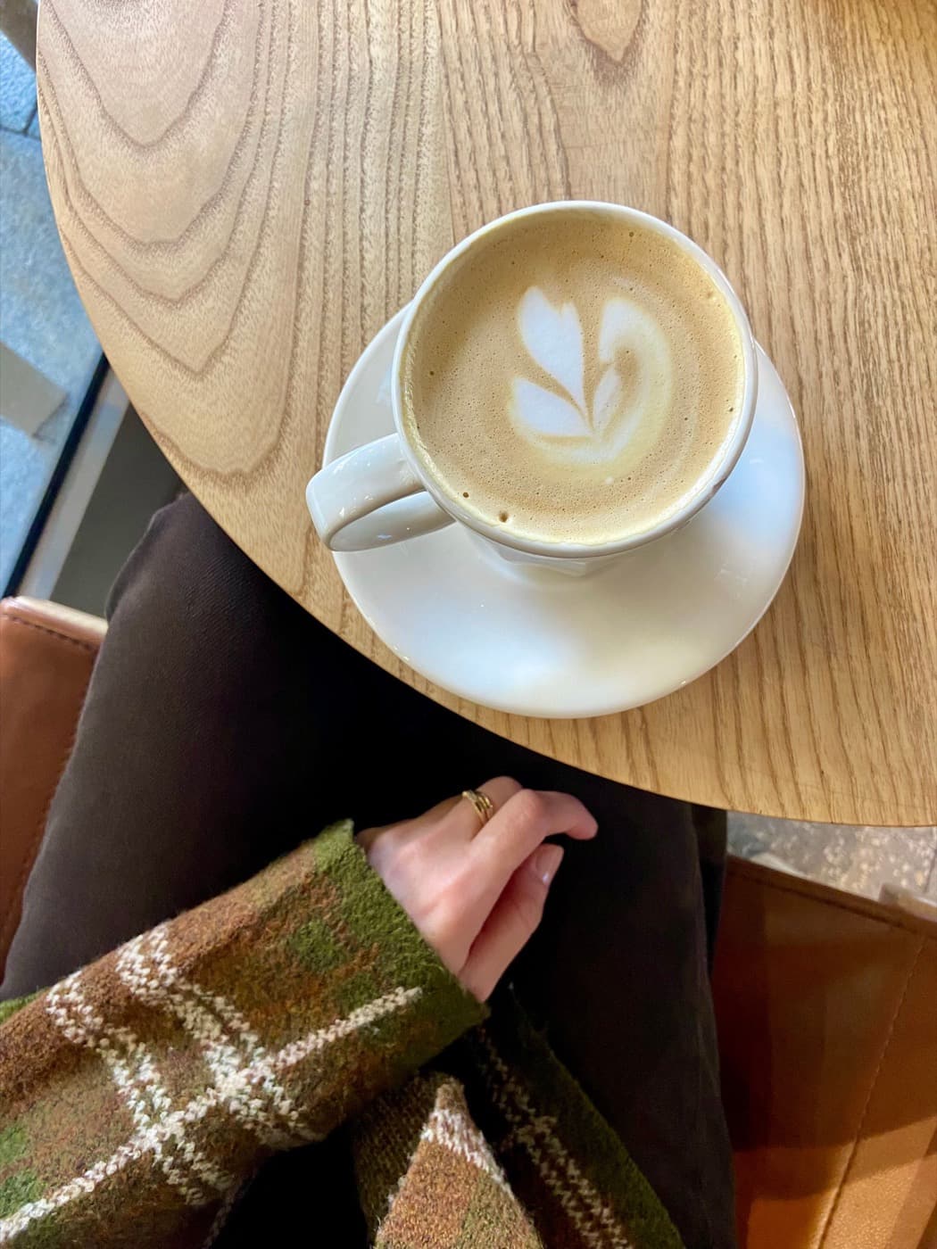 A latte on a round oak table beside the plaid wool sleeve of a fall jacket
