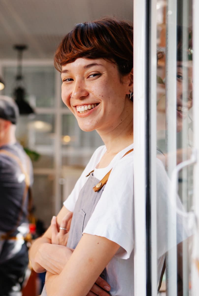 June Park smiling in a canvas apron, leaning in the doorway of the café