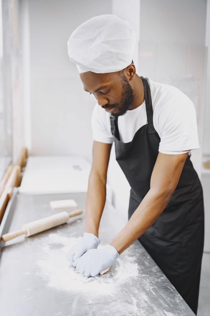 Marcus Bell in a baker’s cap and apron, working dough on a floured steel bench