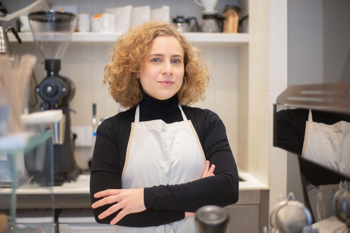 Nora Whitcomb in a white apron, arms folded, beside the espresso grinder